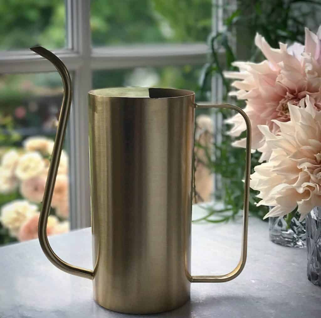 Vattenkannan ELM i mässing från varumärket Nordal, placerad på en bänk framför ett fönster med blommor i bakgrunden.