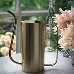 Vattenkannan ELM i mässing från varumärket Nordal, placerad på en bänk framför ett fönster med blommor i bakgrunden.