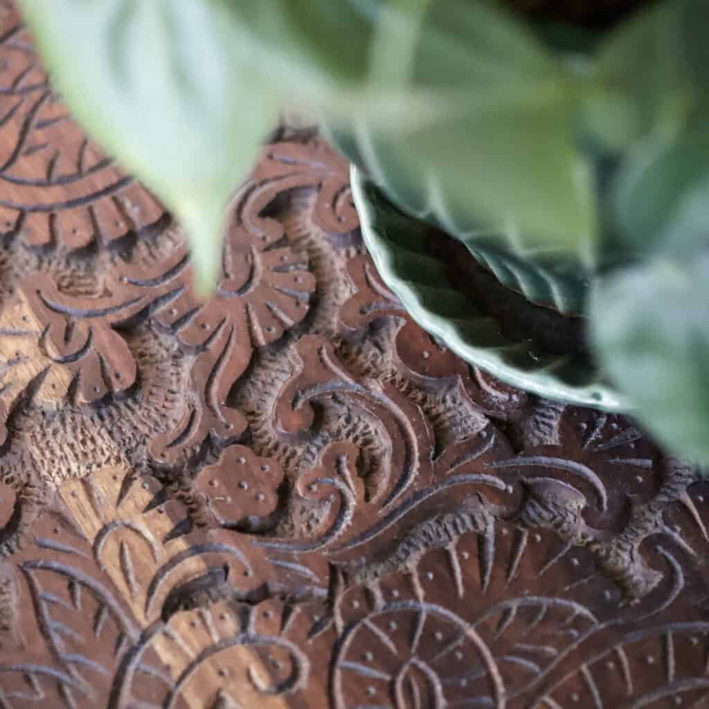 Närbild av ett sidobord i trä från Dutchbone med ett detaljerat och snirkligt mönster på bordsskivan, tillsammans med en grön krukväxt i en keramikskål.