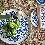 En vacker tallrik med blått mönster, modell MOLLY från Bloomingville, dekorerad med gröna paprikor och vitlök. Tallriken är placerad på en flätad bordstablett omgiven av gröna växter.