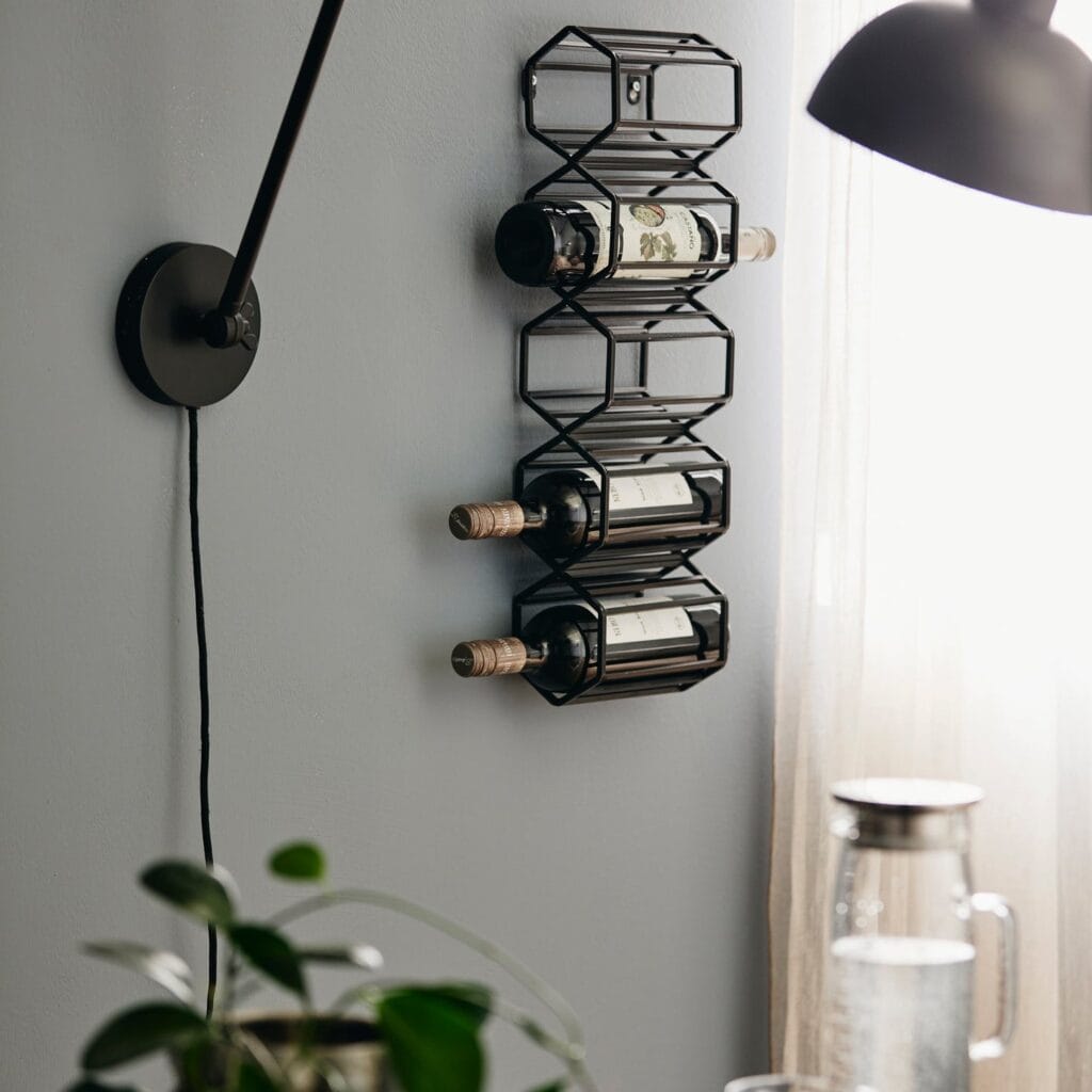Vägghängt svart vinställ från Nordal monterat på en ljusgrå vägg, med tre vinflaskor i förvaring. Stilren och modern design i metall, perfekt för små utrymmen.