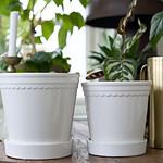 Närbild på två vita krukor i keramik av modellen Astrid från Wikholm Form. Krukorna har en dekorativ vågformad kant och står på matchande fat. De är placerade på ett bord, omgivna av gröna växter och inredningsdetaljer i en hemtrevlig miljö.