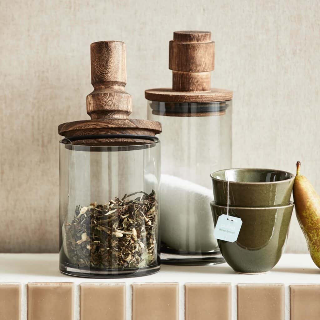Glasburkar Salvie från Nordal med trälock och en grön keramikkopp, presenterade på en köksbänk bredvid en päron, kombinerar både funktionalitet och stil.