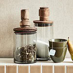 Glasburkar Salvie från Nordal med trälock och en grön keramikkopp, presenterade på en köksbänk bredvid en päron, kombinerar både funktionalitet och stil.