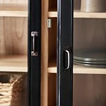 Närbild på handtagen och glasdörrarna på vitrinskåpet VIVA från Nordal, med inblick i trähyllorna och porslin.