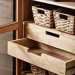 Närbild på en öppen låda och hyllor med flätade korgar i vitrinskåpet VIVA från Nordal, med glasdörrar och trädetaljer.