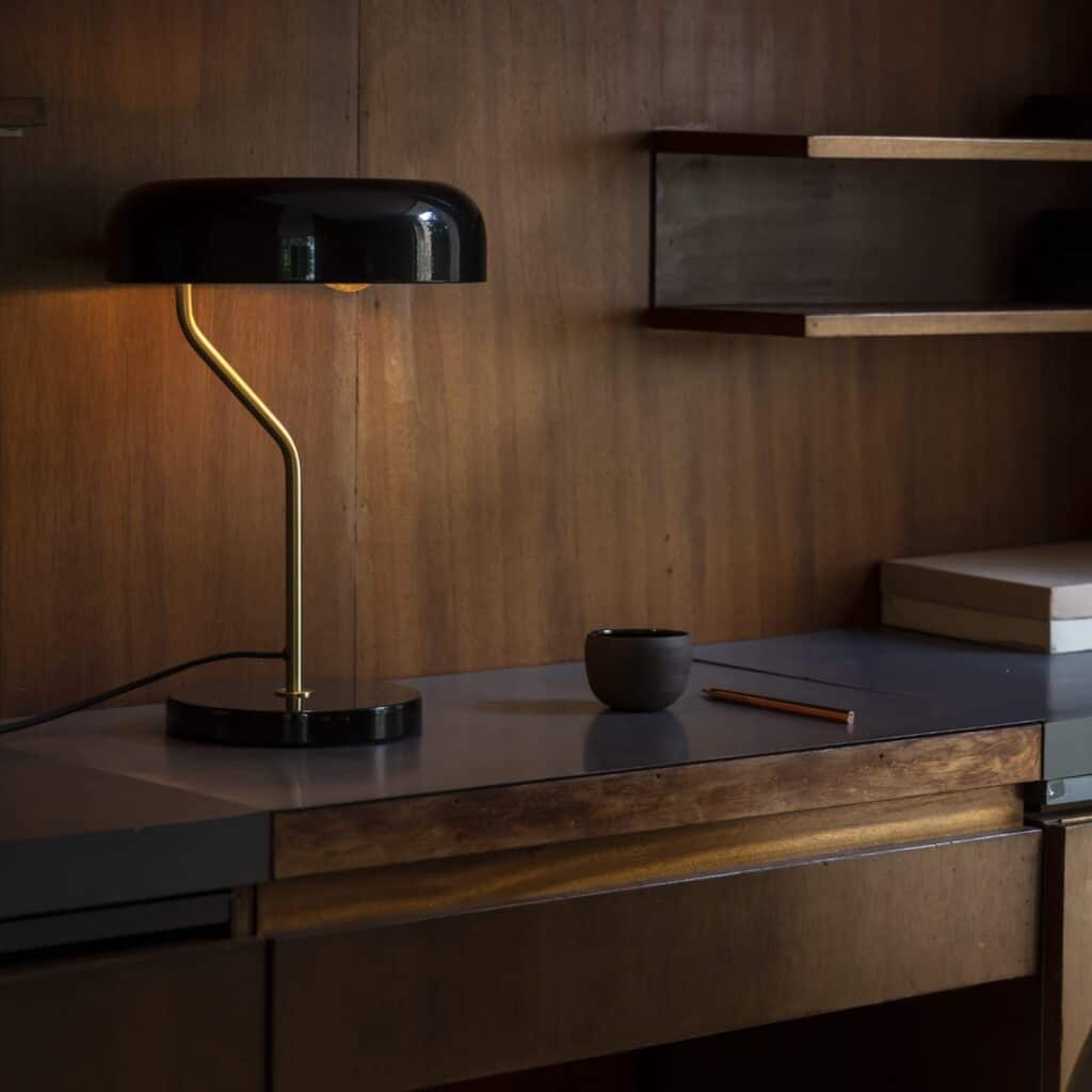 Svart bordslampa med mässingsdetaljer placerad på ett retroinspirerat skrivbord i trä, med en varm och mysig belysning som skapar stämning.