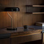 Svart bordslampa med mässingsdetaljer placerad på ett retroinspirerat skrivbord i trä, med en varm och mysig belysning som skapar stämning.