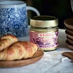 Bild på Gridelli Crema al Pistacchio-siciliansk pistagekräm i en glasburk med en guldlock, omgiven av croissanter på ett träfat och en blåvit mugg i bakgrunden.