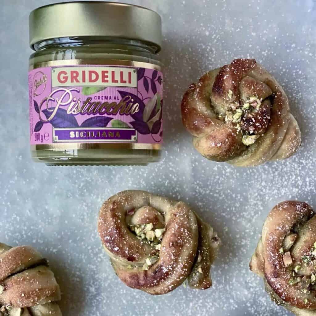Burk med Gridelli Crema al Pistacchio bredvid nybakade pistagefyllda bakverk, pudrade med florsocker.