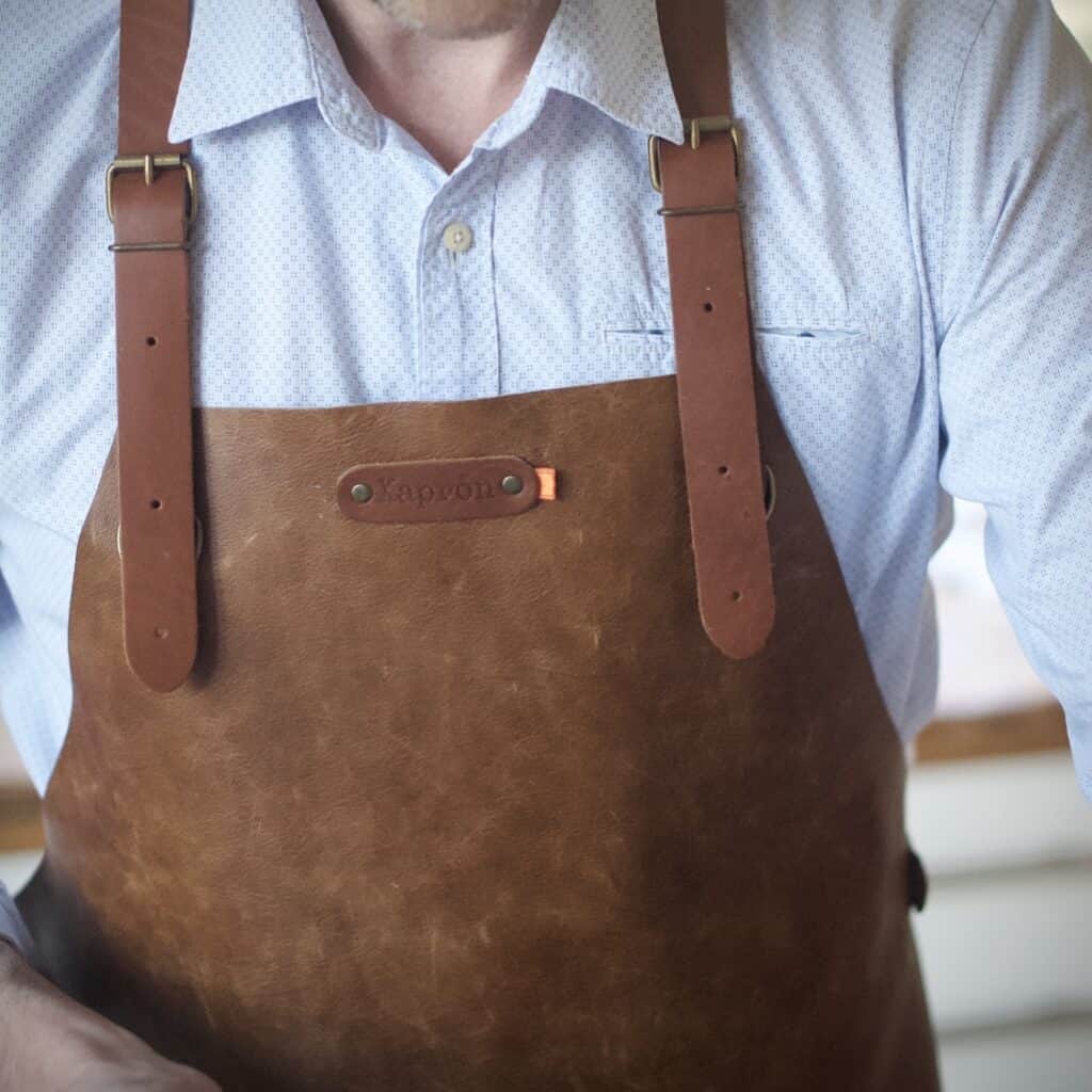 Detaljbild av ett rustikt brunt skinnförkläde UTAH RUST från Xapron, med justerbara läderremmar och snygga metallspännen, buren av en person med en ljus skjorta.