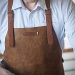 Detaljbild av ett rustikt brunt skinnförkläde UTAH RUST från Xapron, med justerbara läderremmar och snygga metallspännen, buren av en person med en ljus skjorta.