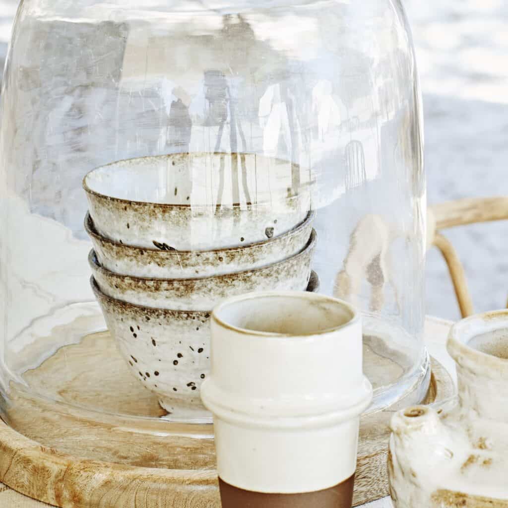 Staplade stengodsskålar med krämvit bas och bruna stänk under en glaskupa, tillsammans med andra rustika keramikföremål på ett träfat.