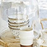 Staplade stengodsskålar med krämvit bas och bruna stänk under en glaskupa, tillsammans med andra rustika keramikföremål på ett träfat.