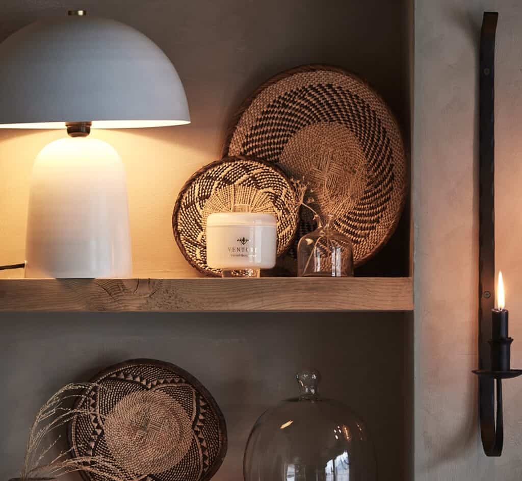 Stilfull inredning med en bordslampa, flätade korgar och väggmonterad ljushållare på en hylla, tillsammans med en glasvas från Affari of Sweden, fylld med dekorativa grässtrån.