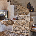 En mysig inredningsmiljö med en beige och svartmönstrad ryamatta draperad över en trappa. Bredvid mattan står tre mönstrade lyktor med tända ljus. På en avsats ligger en kudde med fiskbensmönster och en pläd, tillsammans med en kopp och en skål. Bakom syns en öppen spis och en rustik vägg i jordnära toner.