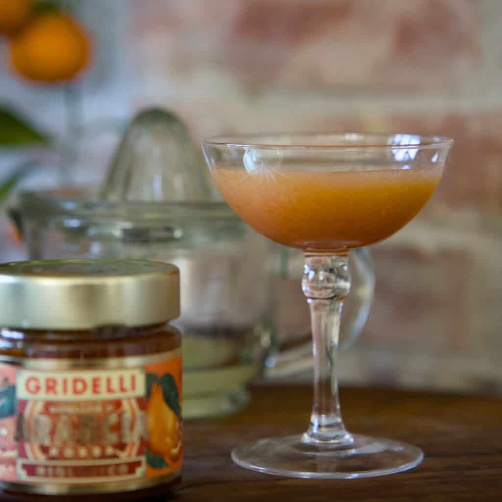 Ett elegant champagneglas vid namn KERSTIN från Strömshaga, fyllt med en fräsch citrusdryck. I bakgrunden syns en glas-citruspress och en burk blodapelsinmarmelad från Gridelli, placerade på ett rustikt träbord mot en tegelvägg.