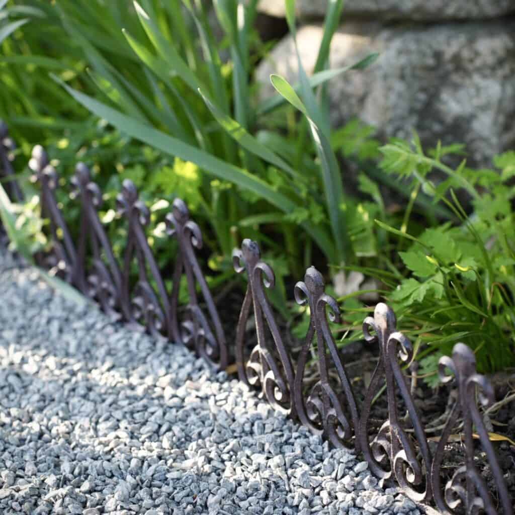 Ett dekorativt kantskydd i gjutjärn från Esschert Design som avgränsar en grusgång från en grönskande rabatt med växter.