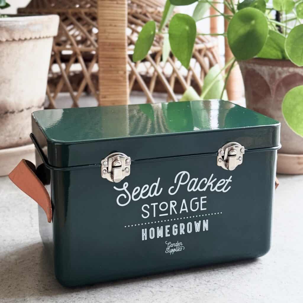 Grön förvaringsburk för fröpåsar från Burgon & Ball med läderhandtag och metallspännen, placerad i en hemtrevlig miljö med gröna växter.