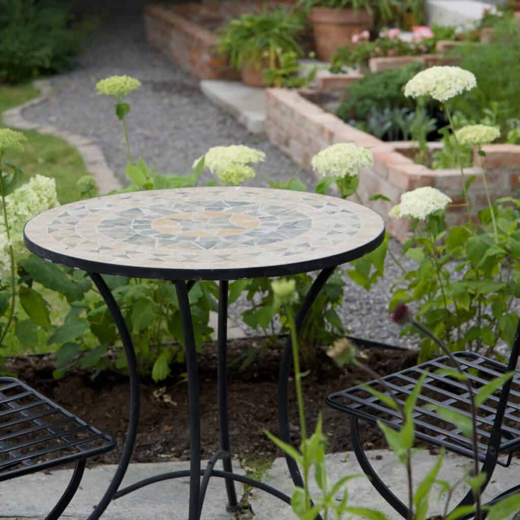 Runt trädgårdsbord i mosaik, modell Venice, med naturfärgade toner och svart smidesunderrede, placerat i en grönskande trädgårdsmiljö.