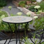 Runt trädgårdsbord i mosaik, modell Venice, med naturfärgade toner och svart smidesunderrede, placerat i en grönskande trädgårdsmiljö.