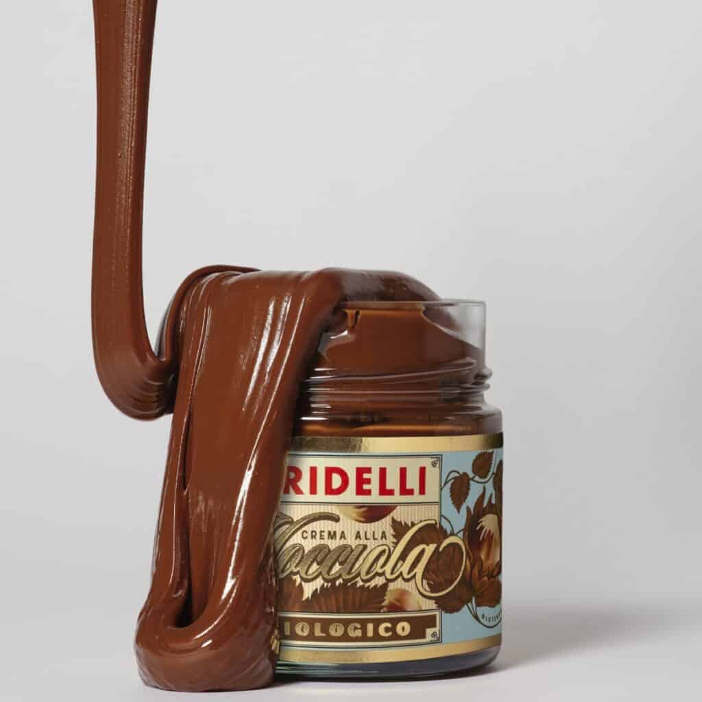 Glasburk med Gridelli Crema alla Nocciola, ekologisk hasselnötskräm, med krämig choklad som rinner över kanten och ner längs burken, fotograferad mot vit bakgrund.