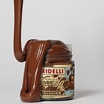 Glasburk med Gridelli Crema alla Nocciola, ekologisk hasselnötskräm, med krämig choklad som rinner över kanten och ner längs burken, fotograferad mot vit bakgrund.