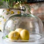 Glaskupa Bella från Affari of Sweden – en elegant, munblåst glaskupa som täcker tre citroner. I bakgrunden syns en stengodskruka med citronmotiv.