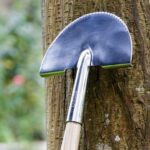 Närbild av Burgon & Balls halvmåneformade kantskärare lutad mot ett träd, med glänsande blad och FSC-certifierat handtag.