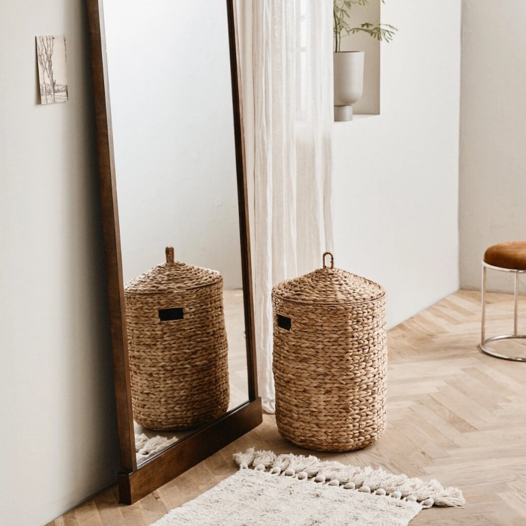 Tvättkorg COSNA i naturfärgad vattenhyacint bredvid en stor träspegel i ett ljust och varmt inrett rum.