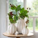 Vit keramikvas LILLIAN i organiskt veckad form, placerad på bord med grönskande björnbärskvistar.