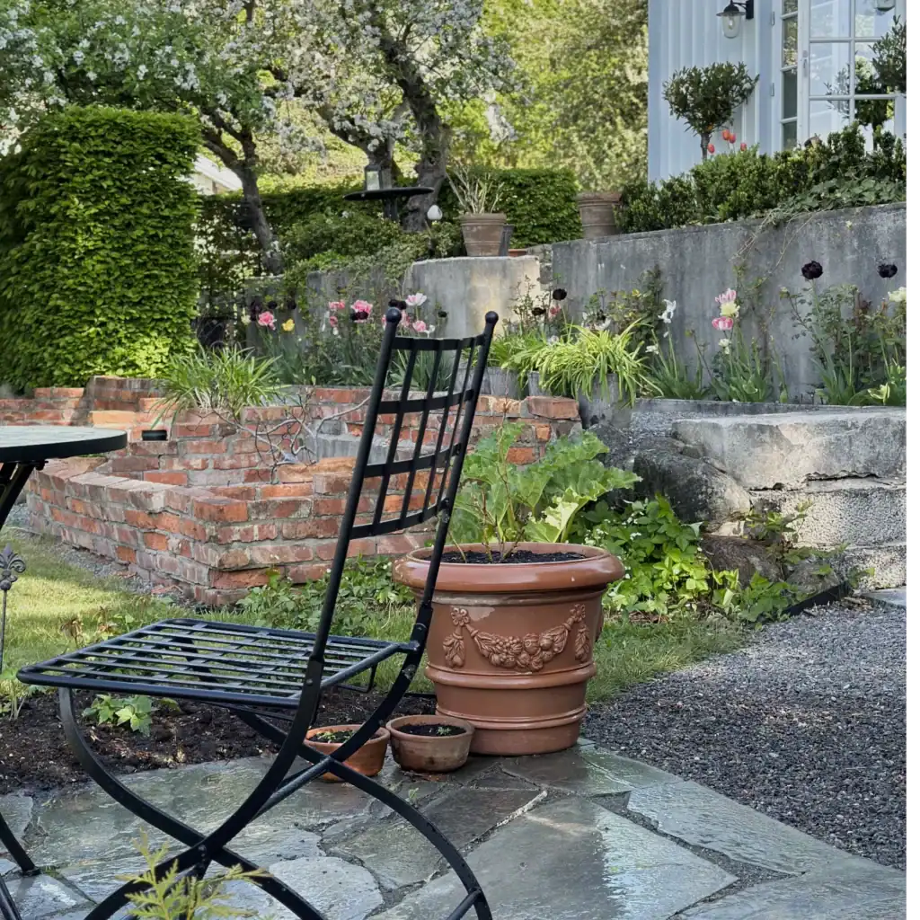 En svart trädgårdsstol vid namn VENICE från varumärket Wikholm form står på en stenlagd uteplats i en lummig trädgård. Bakom stolen syns en terrakottakruka med dekorativa detaljer och en upphöjd rabatt med blommande växter och gröna buskar. I bakgrunden finns ett tegelmurat område och en vit byggnad omgiven av grönska.
