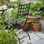 Svart trädgårdsstol i smide, modell Venice, placerad på en stenbelagd uteplats omgiven av gröna växter och terrakottakrukor.