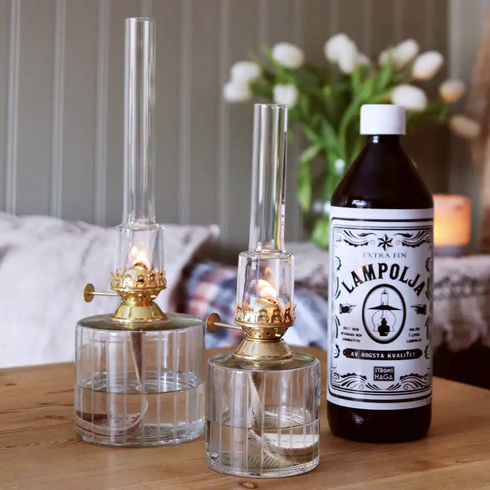 Strömshagas lampolja står bredvid två tända fotogenlampor i glas och mässing på ett träbord.