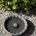 Brunmelerat fågelbad Talia i keramik placeras diskret på en grusbelagd mark vid sidan av gröna växter.