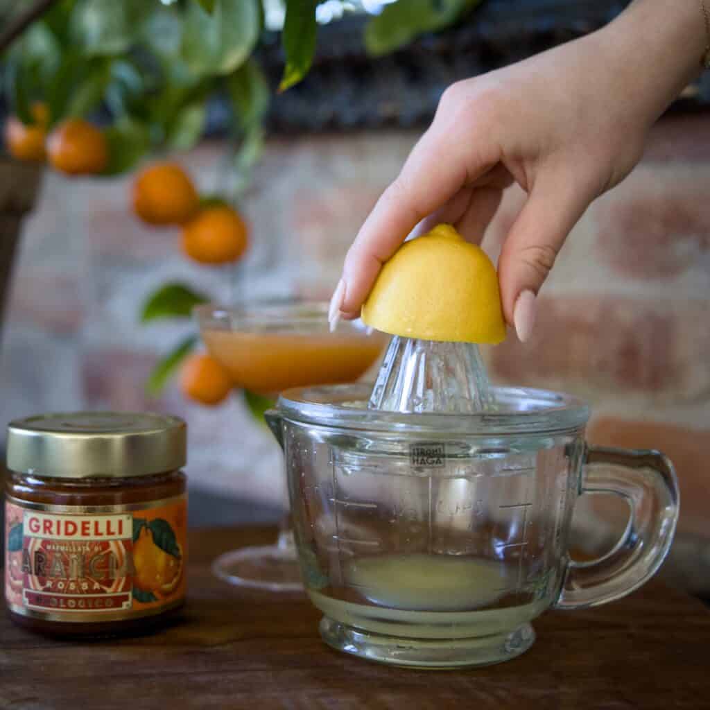 En hand pressar en citronhalva på en klassisk citruspress i glas från Strömshaga. I bakgrunden syns en burk apelsinmarmelad, ett cocktailglas med citrusjuice och en apelsinväxt med mogna frukter mot en tegelvägg.