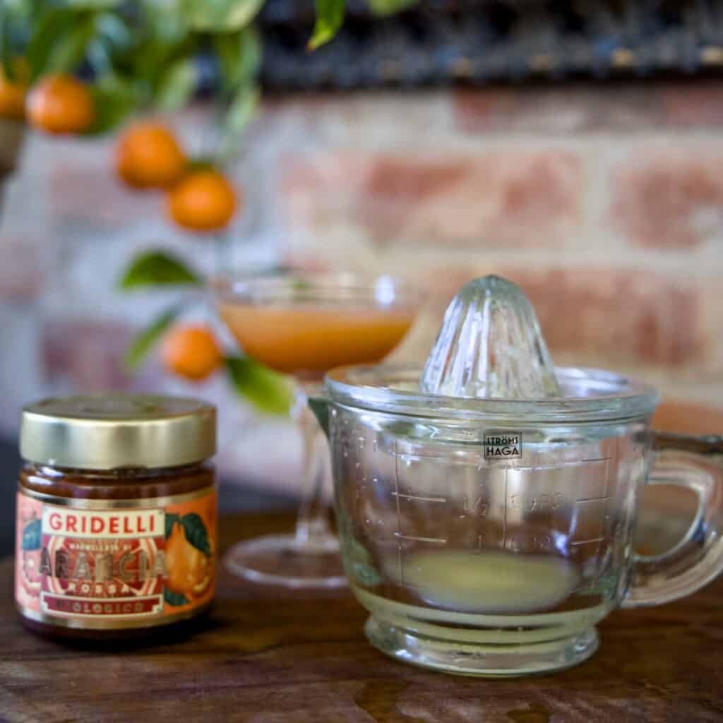 En vacker citruspress i glas från Strömshaga, placerad på ett rustikt träbord tillsammans med en burk apelsinmarmelad och ett cocktailglas fyllt med citrusjuice. I bakgrunden syns en tegelvägg och en apelsinväxt med mogna frukter.