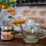 En vacker citruspress i glas från Strömshaga, placerad på ett rustikt träbord tillsammans med en burk apelsinmarmelad och ett cocktailglas fyllt med citrusjuice. I bakgrunden syns en tegelvägg och en apelsinväxt med mogna frukter.