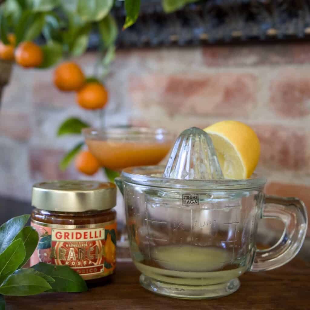 En klassisk citruspress i glas från Strömshaga med en halvpressad citron på toppen. I bakgrunden syns en burk apelsinmarmelad, ett cocktailglas med citrusjuice och en apelsinväxt mot en tegelvägg.