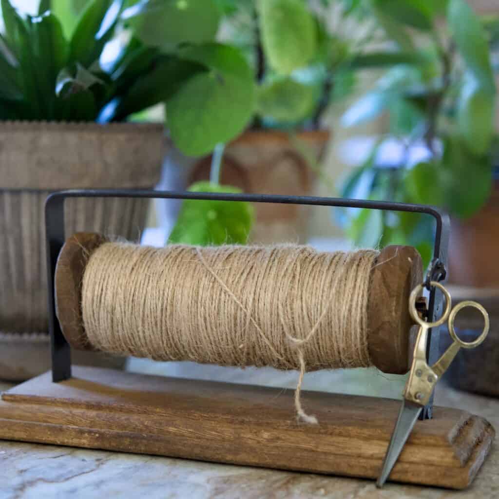 Rustik hållare i trä och metall med jutesnöre och en liten sax, perfekt för pyssel och trädgårdsarbete. Placeras på ett bord med gröna växter i bakgrunden.