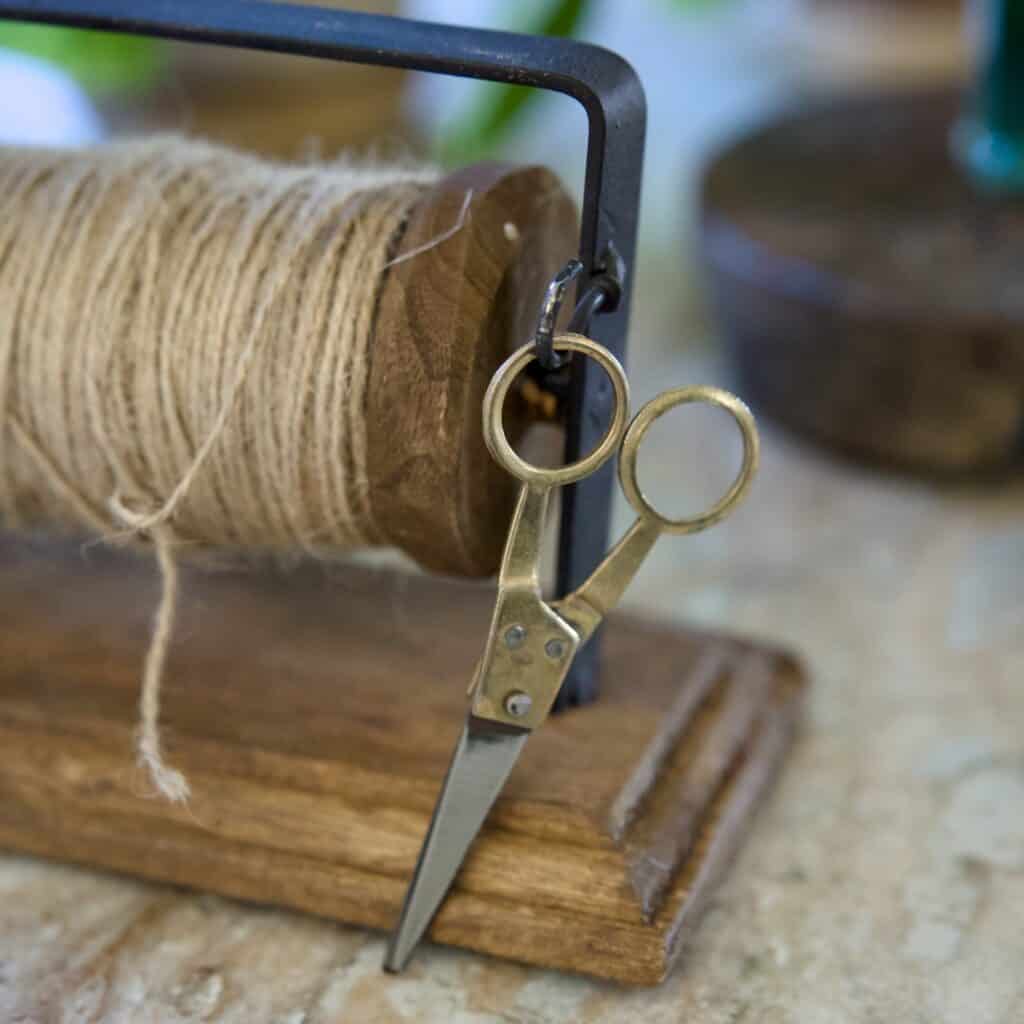 Närbild på en vintageinspirerad sax i metall som hänger på en ställning med jutesnöre, monterad på en träbas. Perfekt för pyssel och dekoration.