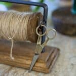 Närbild på en vintageinspirerad sax i metall som hänger på en ställning med jutesnöre, monterad på en träbas. Perfekt för pyssel och dekoration.