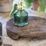 Rustik rund bricka i återvunnet trä med naturlig patina, placerad på en marmoryta. På brickan står en grön glasblomspruta med gulddetaljer.