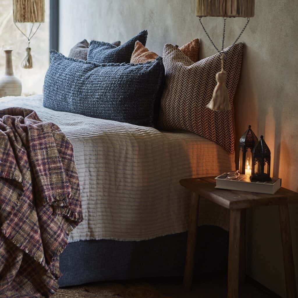 En mysigt inredd sovrumsmiljö med säng klädd i mjuka textilier och kuddar i jordnära toner. På nattduksbordet står lyktan GIZA från Affari of Sweden, tänd för att skapa en varm och stämningsfull atmosfär.