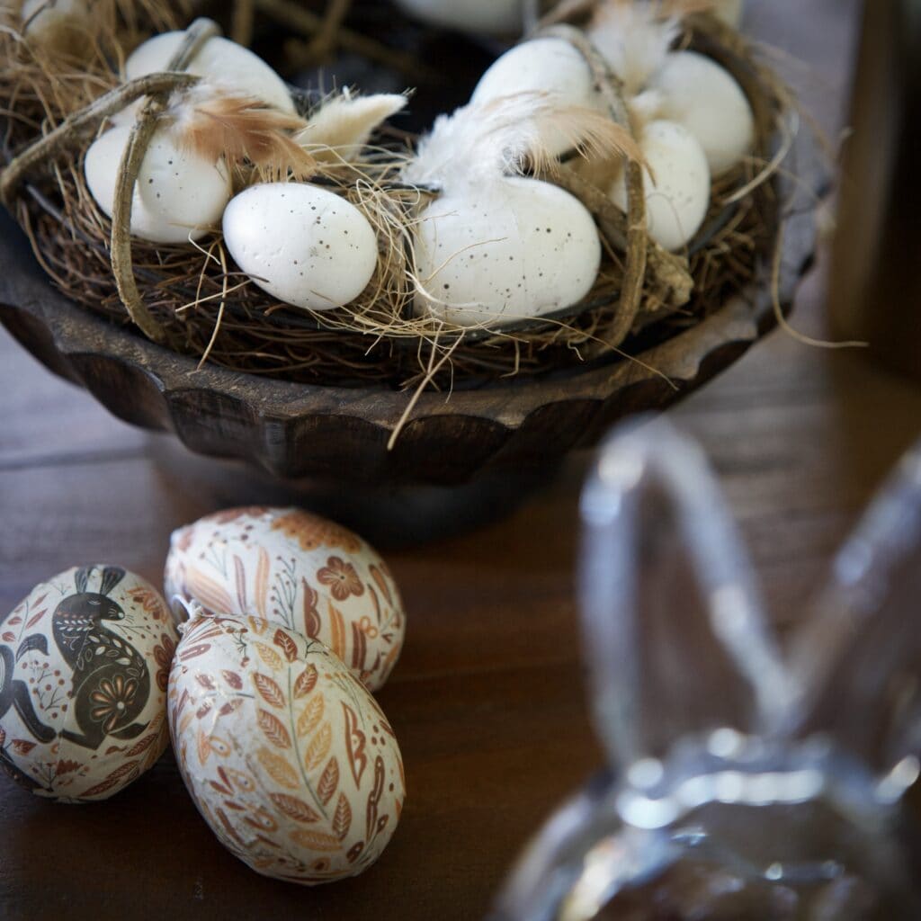 Dekorationskrans med vita ägg och fjädrar i naturmaterial, tillsammans med tre handmålade påskägg i papper i varma toner, liggande på ett träbord.