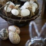 Dekorationskrans med vita ägg och fjädrar i naturmaterial, tillsammans med tre handmålade påskägg i papper i varma toner, liggande på ett träbord.