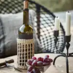SUNNY vinflaskhållare från Affari of Sweden på ett träbord, stylad med en vinflaska, druvor och en rustik ljusstake i en mysig utomhusmiljö.