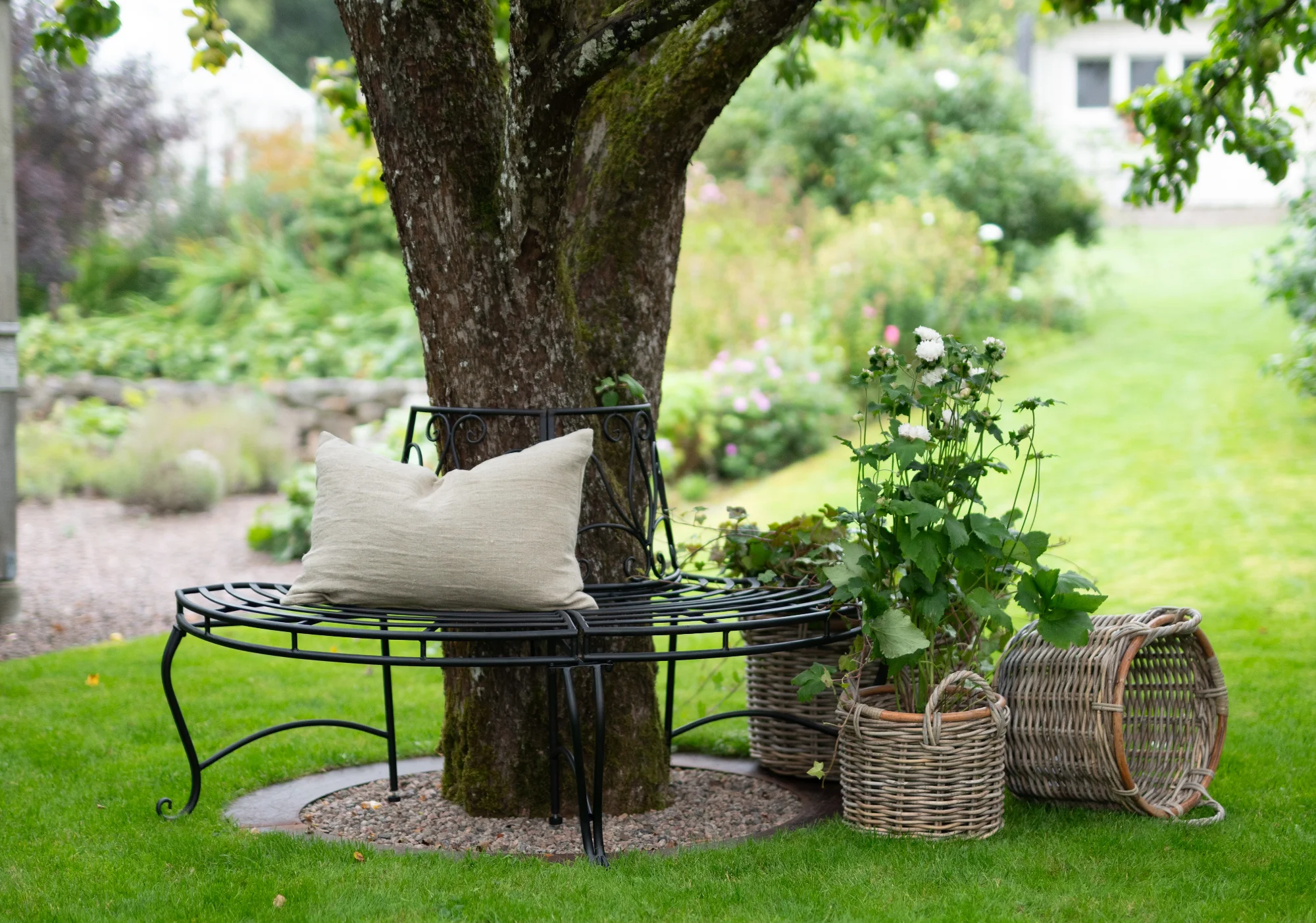 Stilfull trädgårdsmöblering med bekväm bänk under ett frodigt träd, omgivet av grönskande växter och rustika korgar, inspirerar till 'Skapa din drömträdgård'.
