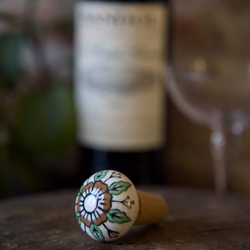 Handmålad vinkork från Madam Stoltz i stengods och kork, med ett vackert blommönster i grönt och brunt. I bakgrunden syns en vinflaska och ett glas, vilket skapar en elegant och stämningsfull miljö.