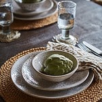 En dukning med tallrikar och skålar från serien Annabelle av Affari of Sweden, med beige prickigt mönster, placerade på runda bordstabletter av flätad halm. En skål innehåller en kronärtskocka, och bredvid tallrikarna ligger en mönstrad servett. Glas med vatten och bestick är också placerade på bordet.